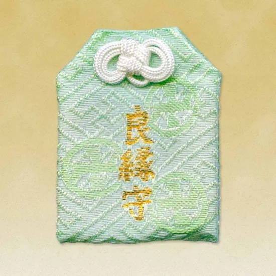 A light green omamori charm with a gold-colored kanji inscription and a white decorative knot. The fabric has a subtle woven pattern.