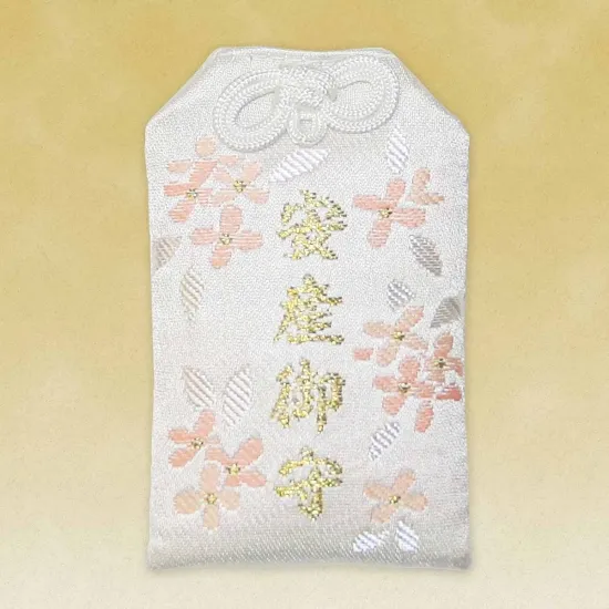 A white omamori charm with a decorative white knot at the top. The fabric is decorated with pink and gold embroidered cherry blossoms and gold kanji characters.