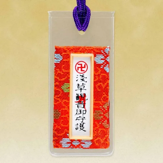 A small, rectangular Japanese omamori charm in a clear plastic sleeve. The charm inside features a green fabric with a subtle pattern and a white central panel with black and red Japanese characters. A purple string is attached at the top.