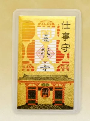 A card-shaped Japanese omamori amulet. It is golden with a detailed illustration of a building and lanterns. Japanese characters are at the top. The amulet is in a clear plastic case.