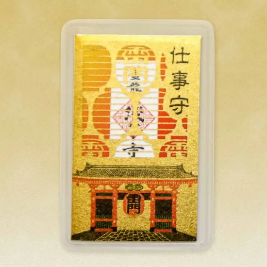 A card-shaped Japanese omamori amulet. It is golden with a detailed illustration of a building and lanterns. Japanese characters are at the top. The amulet is in a clear plastic case.