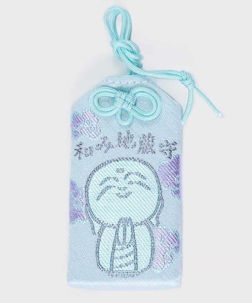 Light blue Japanese omamori with a black embroidered Jizō Bodhisattva figure and purple flowers.