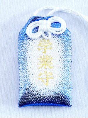 A white, rectangular omamori charm with a dark blue border. The kanji for "Gakugyo Mamori" are written in red in the center. A golden chrysanthemum emblem is at the top