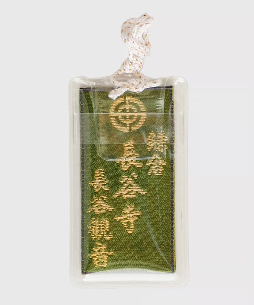 Dark olive green omamori, encased in clear plastic, with gold embroidered text and a circular crest symbol.