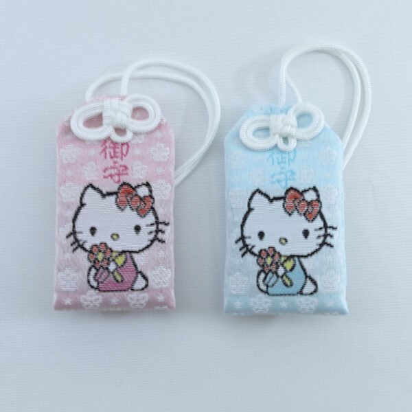 A close-up shot of a small, square, pink & blue omamori amulets featuring a kawaii Hello Kitty face embroidered on the front, with a gold strap.