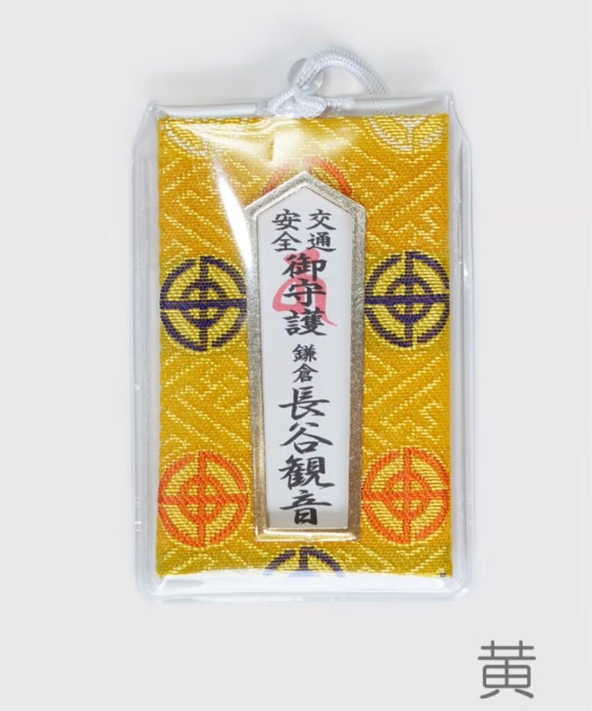 Yellow omamori with a patterned background, encased in clear plastic. A vertical white plaque has black and red Japanese text, including "Traffic Safety" and "Hase-Kannon, Kamakura.