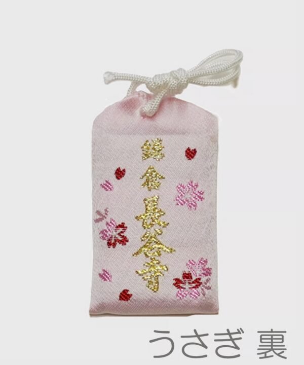 Light pink omamori with small pink cherry blossom designs, tiny red rabbit silhouettes, and gold embroidered Japanese text.