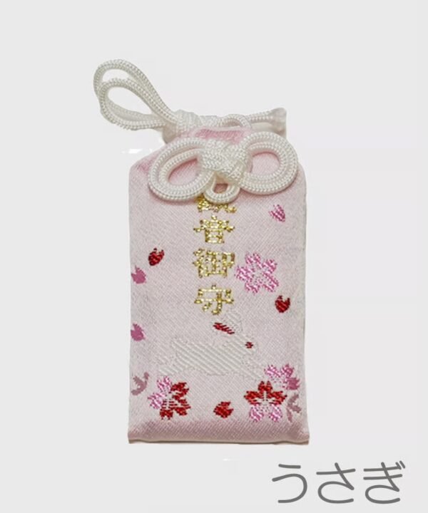 Light pink omamori with small pink cherry blossom designs, tiny red rabbit silhouettes, and gold embroidered Japanese text.