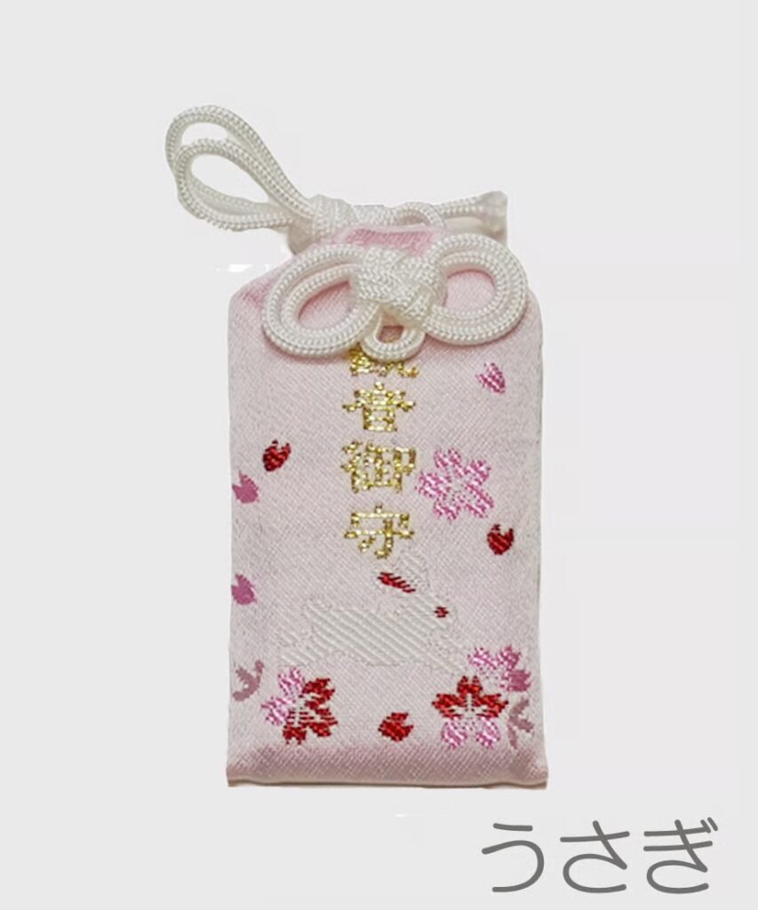 Light pink omamori with small pink cherry blossom designs, tiny red rabbit silhouettes, and gold embroidered Japanese text.