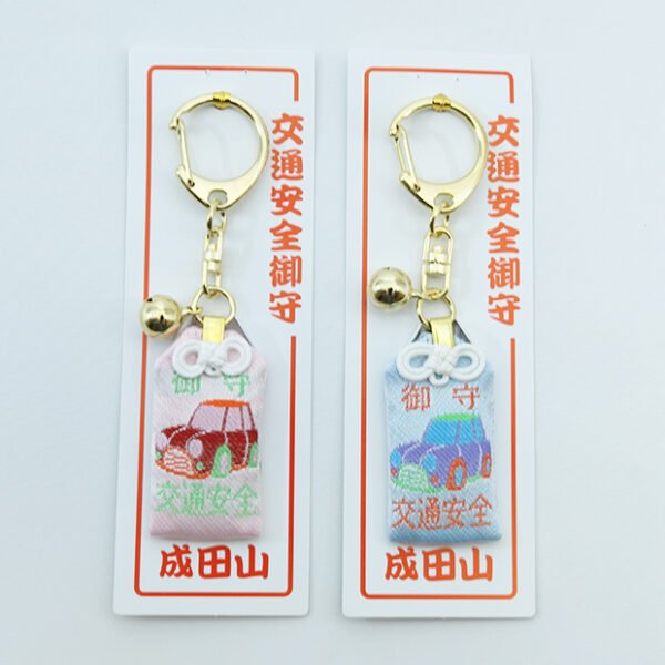 Two Japanese Omamori traffic safety amulets side by side, one is pink and the other is blue, with embroidered car designs and small bells.