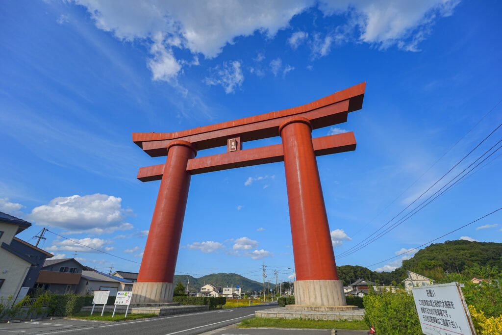 Massive vermilion O-torii gate of Saijo Inari temple, towering over the surrounding landscape and fields under a blue sky.