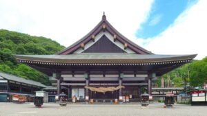 iew of the ornate main hall of Saijo Inari, a large wooden structure with traditional curved roofs and intricate carvings, framed by temple banners and a wide plaza.