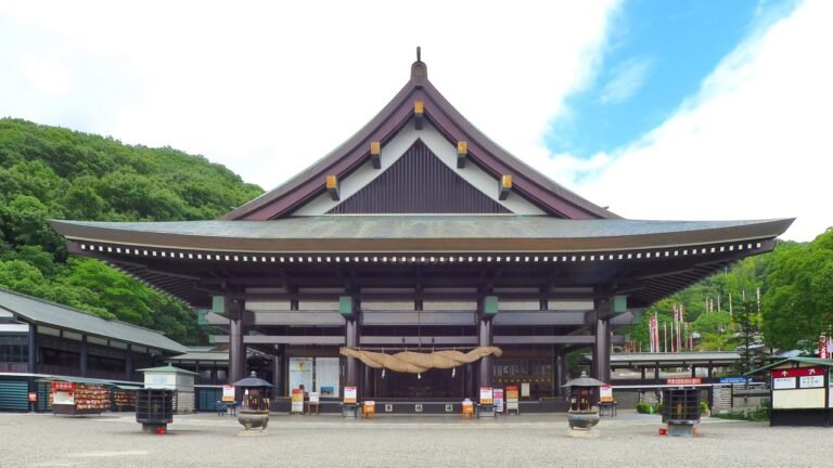 iew of the ornate main hall of Saijo Inari, a large wooden structure with traditional curved roofs and intricate carvings, framed by temple banners and a wide plaza.