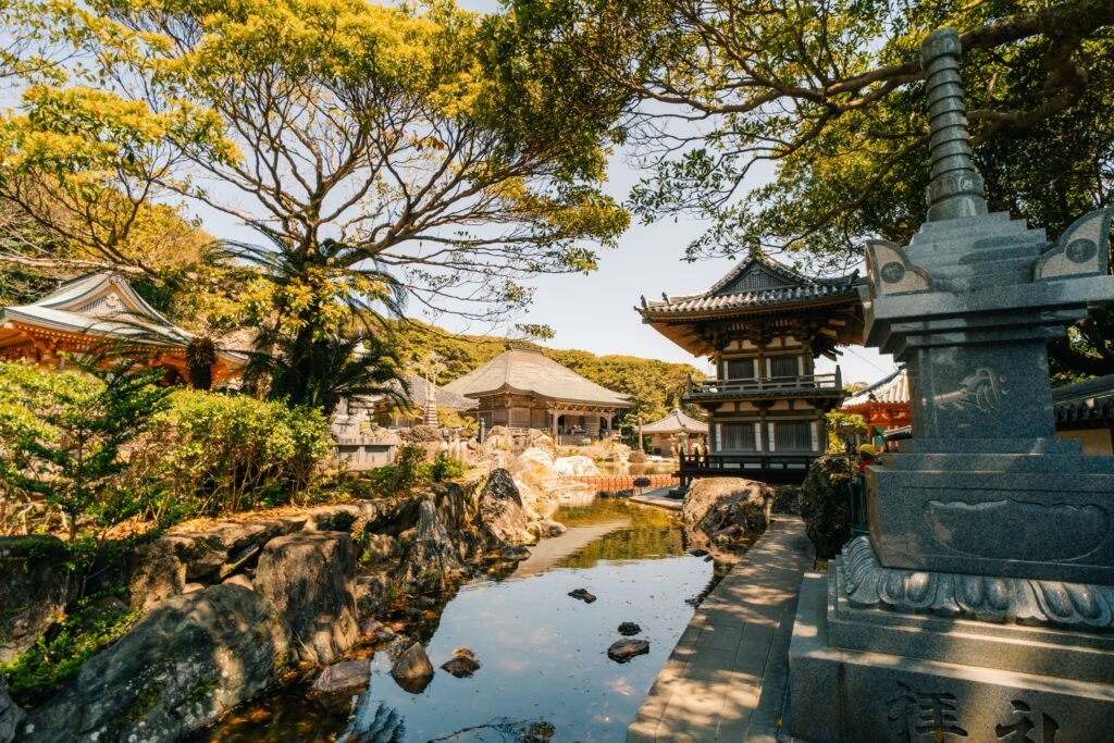 Japanese temple grounds in Shikoku with a clear pond, stone lantern, traditional wooden buildings, a multi-story pagoda, and surrounding green trees on a sunny day.